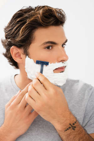 Young man with foam on face shaving with razor isolated on grayの写真素材