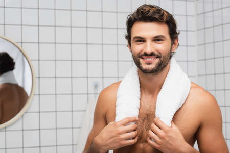 Smiling shirtless man touching towel and looking at camera in bathroomの写真素材
