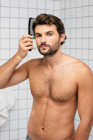 Muscular man combing hair and looking at camera in bathroomの写真素材