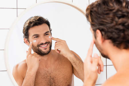 Positive man applying cosmetic cream on face near mirror on blurred foreground in bathroomの写真素材
