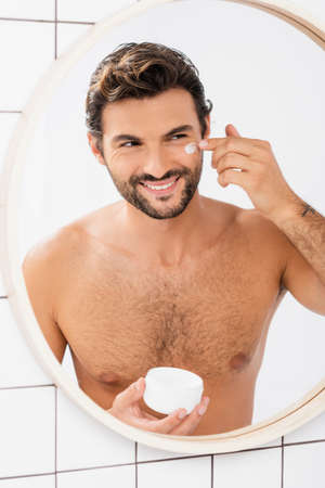 Cheerful man applying face cream while looking at mirror in bathroomの写真素材
