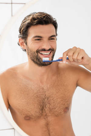 Shirtless man smiling at mirror while holding toothpaste and toothbrush in bathroomの写真素材