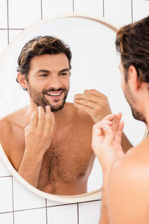 Shirtless man smiling while holding dental floss near mirror on blurred backgroundの写真素材