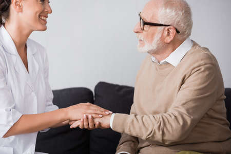 side view of aged man in eyeglasses holding hands with social worker at homeの写真素材