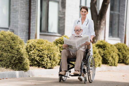cheerful social worker walking with disabled elderly man reading newspaper in wheelchairの写真素材