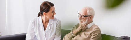 geriatric nurse and aged man talking while sitting on sofa at home, banner