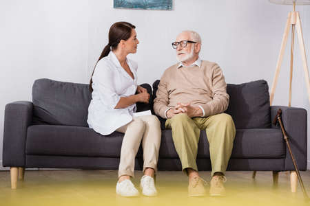 geriatric nurse talking to senior man while sitting on sofa at home on blurred foreground