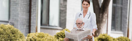 cheerful geriatric nurse walking with handicapped senior man reading newspaper, banner
