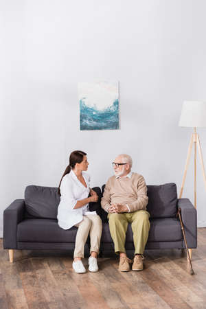 aged man in eyeglasses talking to geriatric nurse while sitting on sofa at homeの写真素材