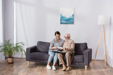 elderly father with adult daughter browsing photo album while sitting on sofa at homeの写真素材