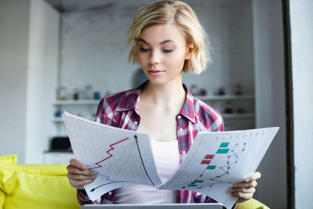 blonde woman in checkered shirt working from home and looking at graphsの写真素材