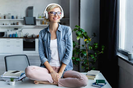 young blonde woman in eyeglasses listening music and sitting on tableの写真素材