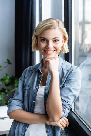 portrait of smiling blonde woman standing near window
