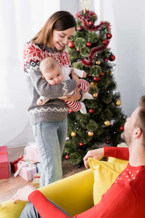smiling woman holding in arms infant boy near husband and christmas tree on blurred background
