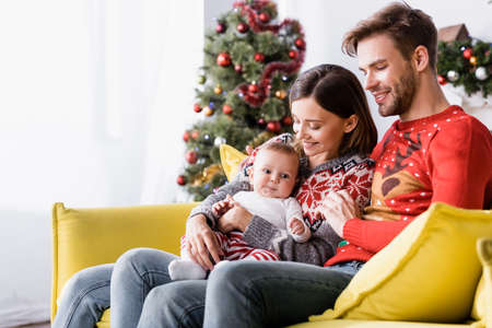 happy woman holding in arms baby boy near cheerful husband near blurred christmas tree on backgroundの写真素材