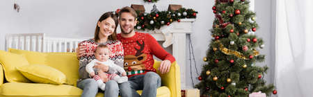 happy family with baby boy sitting on sofa near christmas tree, banner