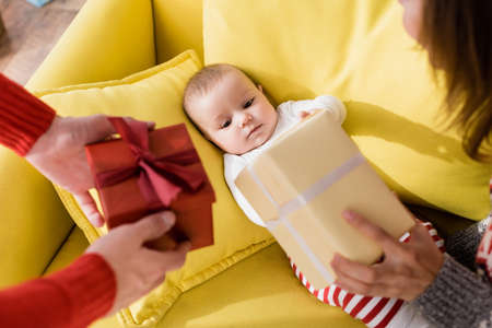 infant son lying on sofa and looking at parents holding wrapped christmas presents on blurred foregroundの写真素材