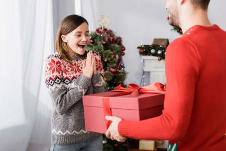 pleased man in red sweater holding wrapped present near excited wife and christmas tree on blurred...