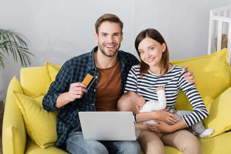 happy man holding credit card near wife with infant son and laptop