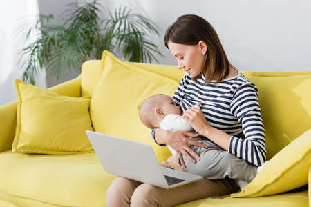 woman holding in arms infant son while breastfeeding near laptop while sitting on sofa