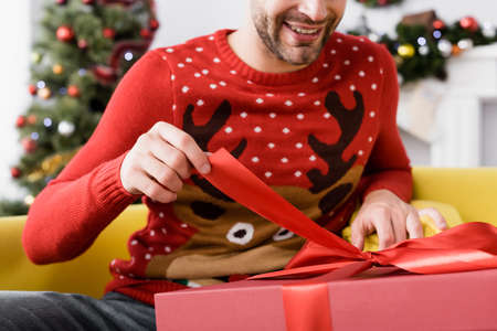 cheerful man in red sweater holding ribbon on present while sitting on sofa