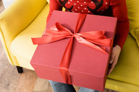 cropped view of man in red sweater holding wrapped present while sitting on sofa with