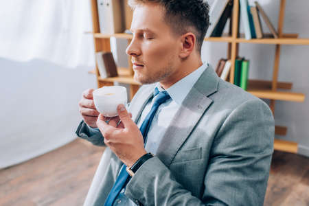 Young businessman with closed eyes holding coffee cup in officeの写真素材