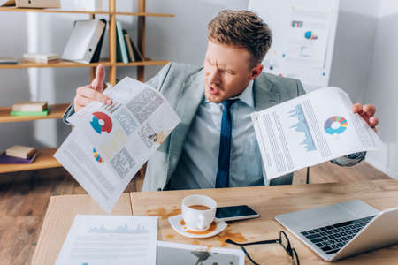 Aggressive businessman holding dirty documents near pouring out coffee and devices in office