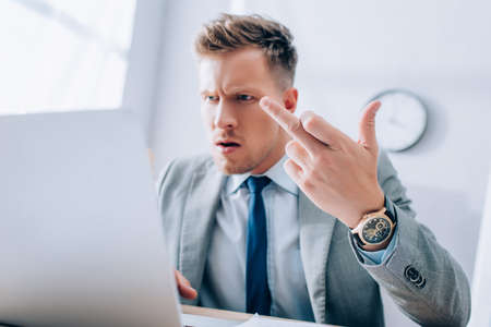Angry businessman showing middle finger while using laptop on blurred foreground in officeの写真素材