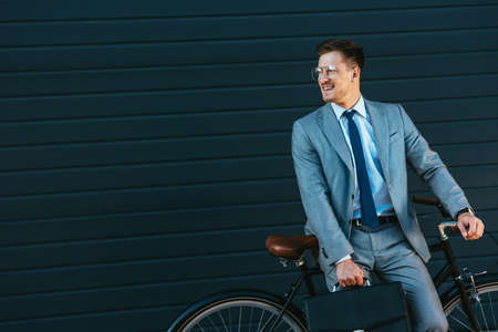 Smiling businessman holding briefcase near bicycle and buildingの写真素材