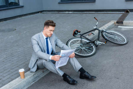 Businessman reading newspaper near coffee to go and bike on walkwayの写真素材