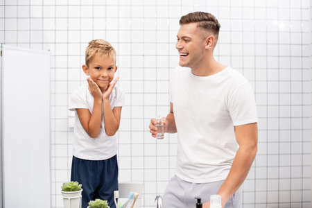 Cheerful father with shaving lotion laughing, while looking at son with hands near cheeks standing on chair in bathroomの写真素材