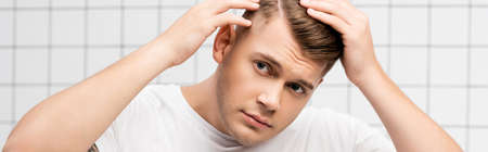 Worried young adult man checking hair in bathroom, banner