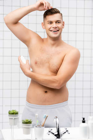 Smiling shirtless man using deodorant while standing near sink with toiletries in bathroomの写真素材