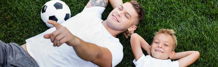 Overhead view of smiling son looking at camera while lying near father pointing with finger on grass, bannerの写真素材
