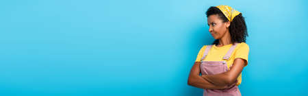 skeptical african american woman standing with crossed arms and looking away on blue, bannerの写真素材