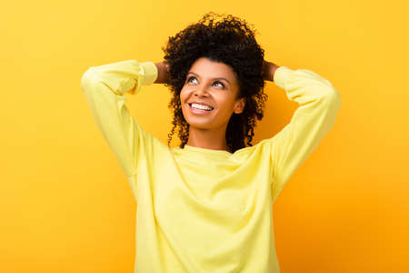 happy african american woman looking away while fixing curly hair on yellowの写真素材