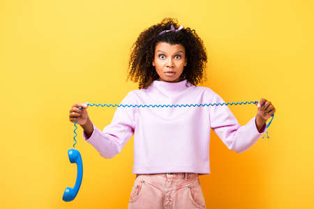 shocked african american woman holding blue retro phone on yellow