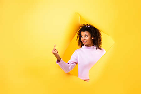 happy african american woman showing middle finger through hole in ripped paper on yellow background