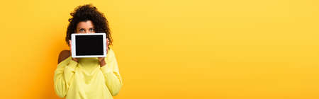 african american woman covering face with digital tablet with blank screen on yellow, banner