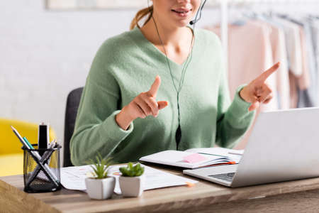 Cropped view of freelancer in headset pointing with fingers during video call on laptop on blurred...