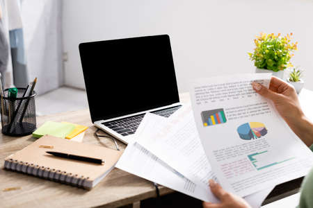 Cropped view of freelancer holding papers with charts near laptop with blank screen and notebook on...