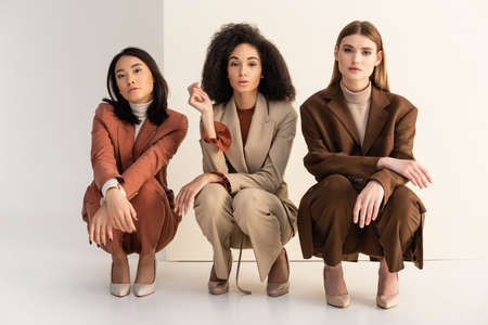 interracial young women in trendy suits sitting and posing on whiteの写真素材