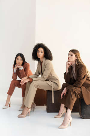 full length of multicultural women in trendy formal wear sitting on suitcases on whiteの写真素材