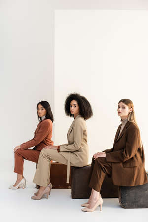 full length of multicultural women in trendy suits sitting on baggage on whiteの写真素材