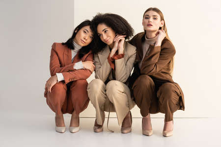 young interracial women in trendy suits sitting and posing on whiteの写真素材