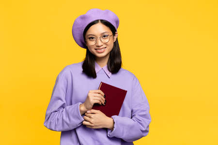 happy asian woman in beret and eyeglasses holding notebook isolated on yellowの写真素材