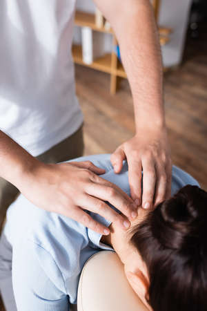 Cropped view of masseur doing seated massage of neck for brunette woman with blurred office on backgroundの写真素材