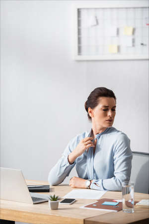 Businesswoman with painful neck untying blouse, while sitting at workplace on blurred backgroundの写真素材