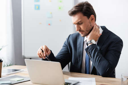 Businessman with painful neck sitting at workplace on blurred backgroundの写真素材